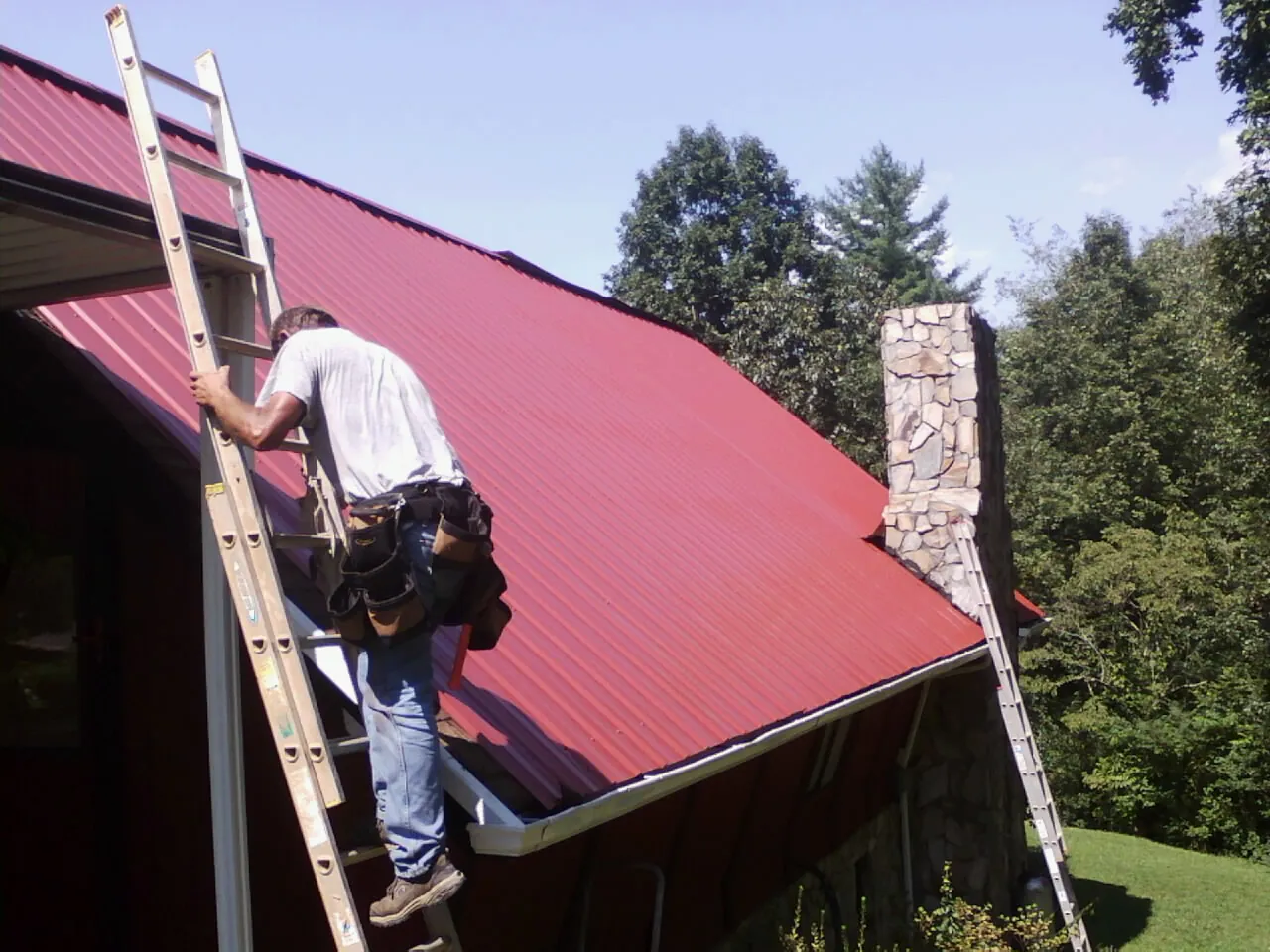 Roofer on a ladder beside a red metal roof and stone chimney — Soffit & Fascia Repair by Roofing Brothers in Martell